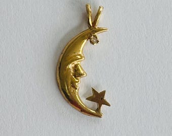 May include: A gold-colored pendant featuring a crescent moon with a smiling face and a small star. The pendant has a small, clear gemstone near the top. The design is simple and elegant.
