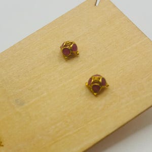 May include: A pair of gold-colored stud earrings with small, round, red gemstones. The earrings are displayed on a light-colored wooden card with the text "FORT KNOX Alameda, CA" printed on it.