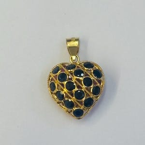 May include: A gold heart-shaped pendant featuring a lattice design with small, round, dark blue gemstones set into each opening. The pendant includes a small gold bail at the top for attachment to a chain, showcasing a detailed metalwork frame.