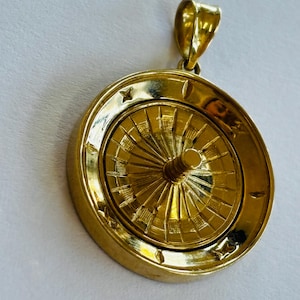 May include: A circular gold pendant designed to resemble a roulette wheel. The piece features a central spinning element with radial spokes and a raised outer rim decorated with small star-shaped engravings. A polished gold bail is attached at the top.