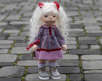 May include: A handmade doll with long white hair and red bows. The doll wears a plaid dress with a red collar and a pink skirt, gray socks, and gray shoes. It holds a small bell. The doll is standing on a brick walkway.