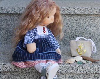 May include: A handmade doll with long, wavy blonde hair, wearing a blue and white checkered dress with a blue striped skirt and white sneakers. A small beige tote bag with a gold anchor design sits nearby, along with seashells and small stones.