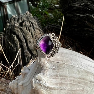 May include: A silver ring with a large, faceted amethyst gemstone surrounded by a floral design. The ring is set on a piece of driftwood.