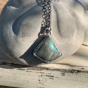 May include: A silver pendant necklace with a turquoise stone set in a geometric shape. The stone has a green and blue color with a marbled pattern.
