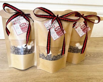 May include: Three brown paper bags filled with hot cocoa mix ingredients. Each bag has a red and black checkered ribbon tied around it and a red and white tag that says "Homemade Hot Cocoa Mix". The bags are filled with chocolate chips, mini marshmallows, and brown sugar.