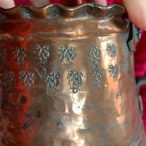 以下が含まれることがあります： 波打つ縁取りと浮き彫りの花柄が施された銅製の鍋。鍋には取っ手が付いており、ハンマーで叩かれた金属でできています。