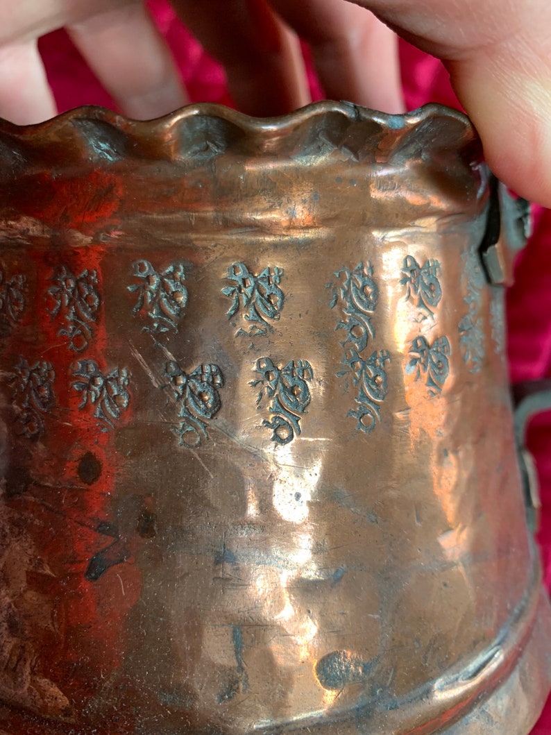 以下が含まれることがあります： 波打つ縁取りと浮き彫りの花柄が施された銅製の鍋。鍋には取っ手が付いており、ハンマーで叩かれた金属でできています。