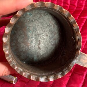 以下が含まれることがあります： 波打つ縁と取っ手のある銅製の鍋。鍋の内側はくすんでいて、緑青がついています。