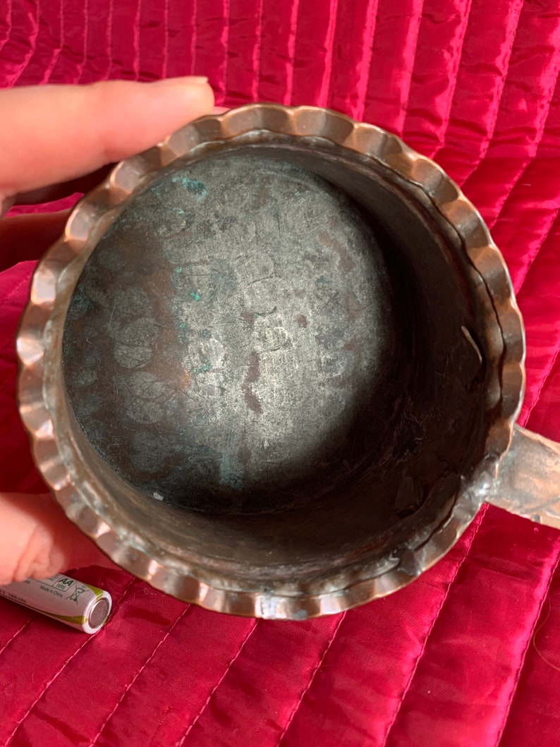 以下が含まれることがあります： 波打つ縁と取っ手のある銅製の鍋。鍋の内側はくすんでいて、緑青がついています。