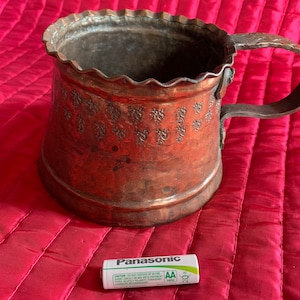 以下が含まれることがあります： 装飾的なエンボス加工のデザインとハンドルが付いた銅製のマグカップ。マグカップは赤いキルティングの表面の上に置かれています。パナソニックの単三電池が手前にあります。