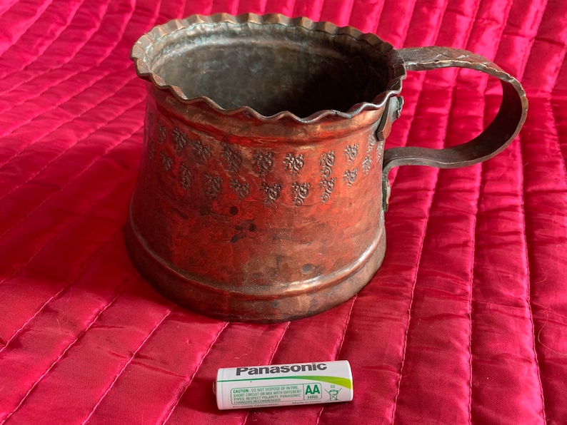 以下が含まれることがあります： 装飾的なエンボス加工のデザインとハンドルが付いた銅製のマグカップ。マグカップは赤いキルティングの表面の上に置かれています。パナソニックの単三電池が手前にあります。