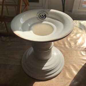 Puede incluir: Un candelabro de cerámica blanca con una base circular ancha y un borde superior acampanado. La superficie superior presenta un logotipo circular negro con el texto G RUSHBROOKE SMITHFIELD y un símbolo R central. Acabado liso y brillante.