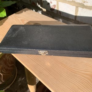 Puede incluir: Un estuche de almacenamiento rectangular de color negro con textura, tapa abatible y un cierre central de metal plateado. La superficie exterior presenta un acabado granulado y los bordes muestran ligeros signos de desgaste.