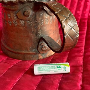 以下が含まれることがあります： ハンマーで叩かれた取っ手と複雑な花柄が施された銅製のマグカップ。マグカップは赤い表面に置かれ、その前にAA電池が置かれている。電池は緑と白で、「CAUTION DO NOT DISPOSE OF IN FIRE, SHORT CIRCUIT OR MIX WITH DIFFERENT TYPES RESPECT POLARITY, PANASONIC CHARGERS RECOMMENDED Panasonic Corporation AA HR6 Made in China EU」という文字が書かれている。