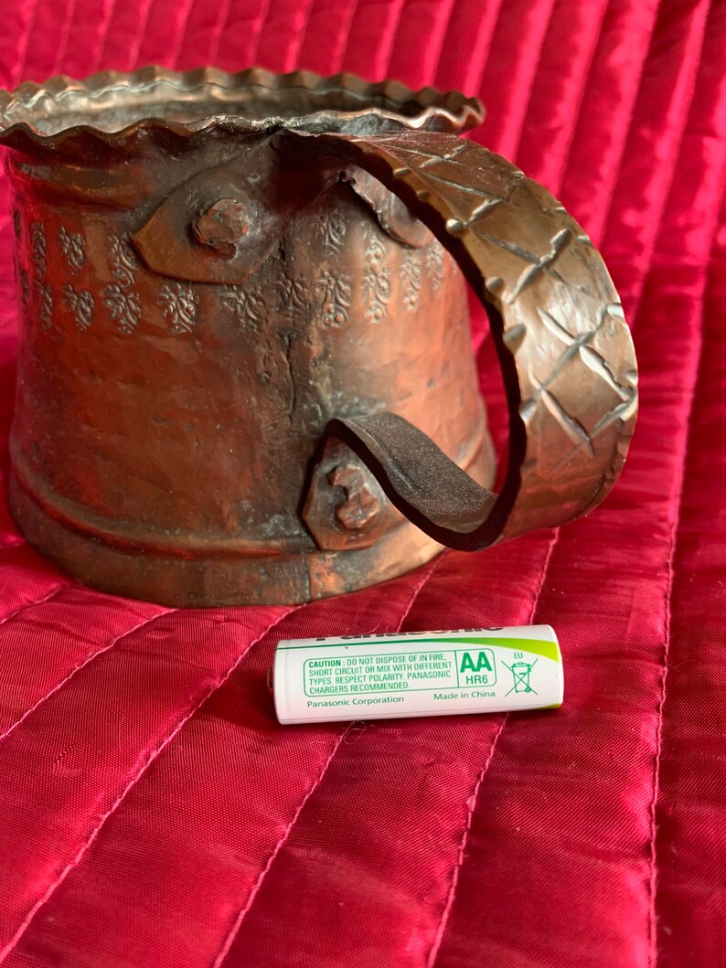 以下が含まれることがあります： ハンマーで叩かれた取っ手と複雑な花柄が施された銅製のマグカップ。マグカップは赤い表面に置かれ、その前にAA電池が置かれている。電池は緑と白で、「CAUTION DO NOT DISPOSE OF IN FIRE, SHORT CIRCUIT OR MIX WITH DIFFERENT TYPES RESPECT POLARITY, PANASONIC CHARGERS RECOMMENDED Panasonic Corporation AA HR6 Made in China EU」という文字が書かれている。