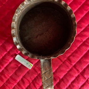 以下が含まれることがあります： 装飾的なハンドルと波状の縁を持つ銅製のマグカップ。マグカップは空で、濃い茶色の緑青がかかっています。