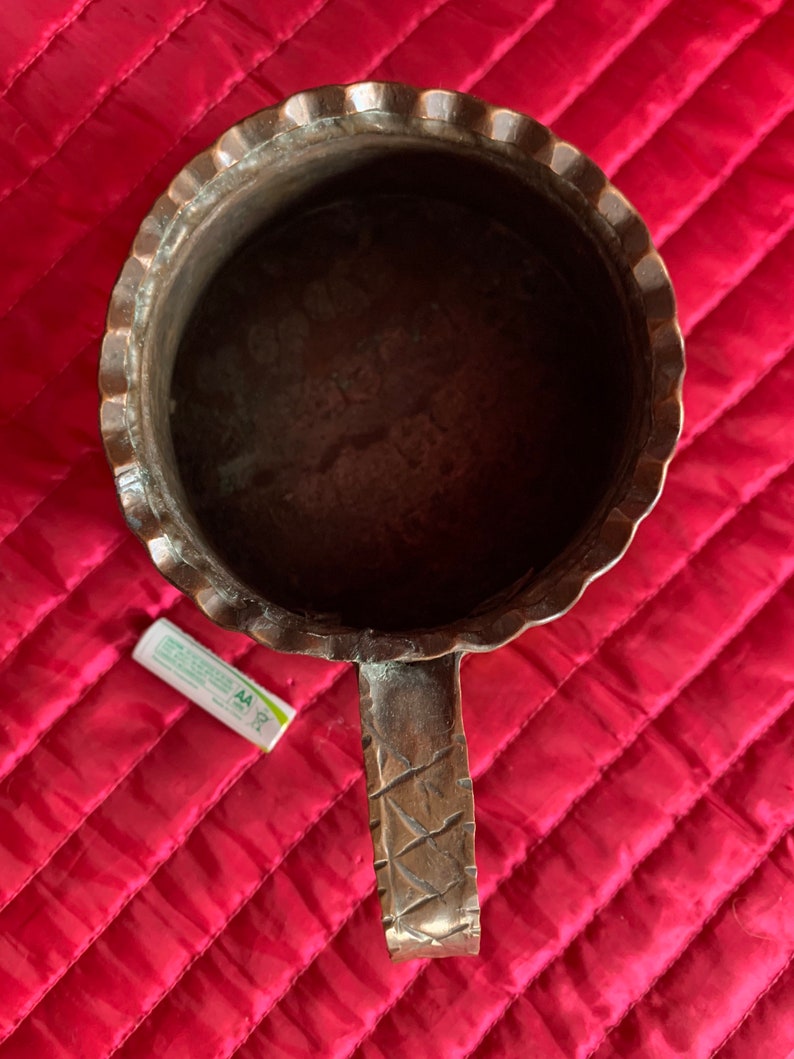 以下が含まれることがあります： 装飾的なハンドルと波状の縁を持つ銅製のマグカップ。マグカップは空で、濃い茶色の緑青がかかっています。