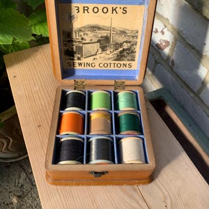 May include: A wooden box containing nine spools of sewing thread in various colours. The interior lid features a label with a black and white illustration of a factory and the text BROOK'S SEWING COTTONS. The spools are arranged in a three-by-three grid.