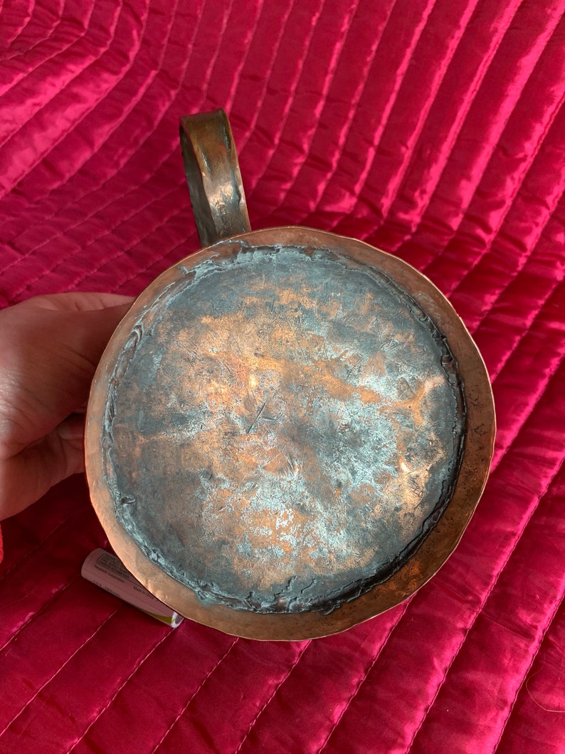 以下が含まれることがあります： 使い込まれた銅製のピッチャー。ピッチャーは、持ち手と丸い本体を持ち、底面が露出しており、銅本来の色合いが見て取れます。