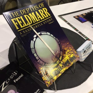 May include: A black book titled "The Depths of Feldmarr: A Saxetertia Story, Volume 1" with a gold banjo on the cover. The book is on a black stand with a black mat underneath. There are business cards stacked on the right side of the image.