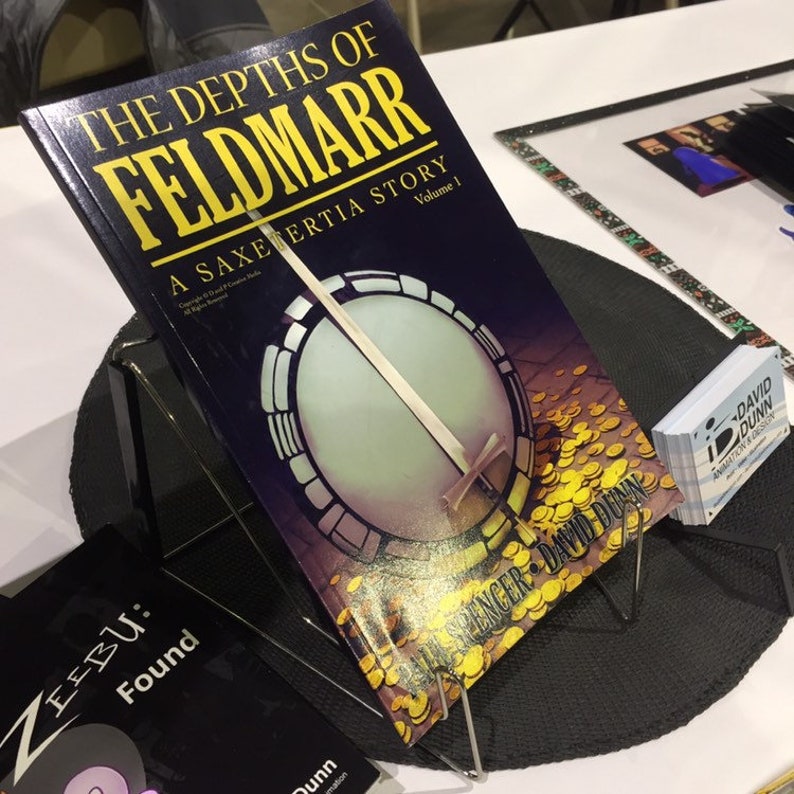 May include: A black book titled "The Depths of Feldmarr: A Saxetertia Story, Volume 1" with a gold banjo on the cover. The book is on a black stand with a black mat underneath. There are business cards stacked on the right side of the image.