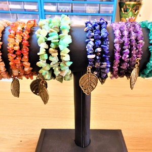 Op de afbeelding: Een display van zes edelsteen chip armbanden in verschillende kleuren: perzik, oranje, groen, blauw, paars en teal. Elke armband heeft een bladbedel. De armbanden worden op een zwarte standaard getoond.