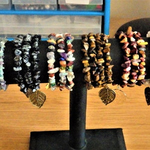 Könnte beinhalten: Eine Auswahl an Armbändern aus Edelstein-Chips in verschiedenen Farben: Grün, Schwarz, Mischfarben, Braun und Weiß. Jedes Armband ist mit einem goldfarbenen Blattanhänger versehen. Die Armbänder werden auf einem schwarzen Ständer vor neutralem Hintergrund präsentiert.
