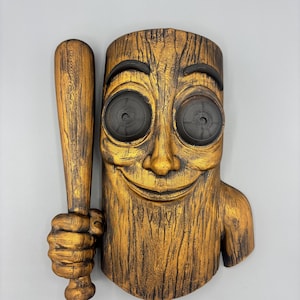 May include: A wall-mounted sculpture featuring a golden-brown, wood-textured figure with a smiling face and large, dark, circular eyes. The figure has a hand gripping a matching golden-brown baseball bat positioned vertically on its left side.