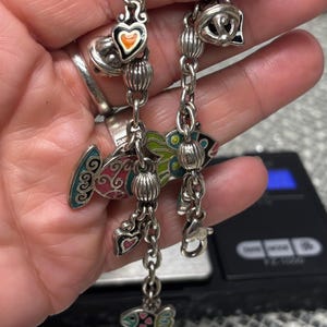 Pu&ograve; includere: Bracciale con ciondoli in argento con pesci in smalto colorato, cuori e perline decorative. Il bracciale ha una chiusura a moschettone ed &egrave; mostrato su uno sfondo sfocato.