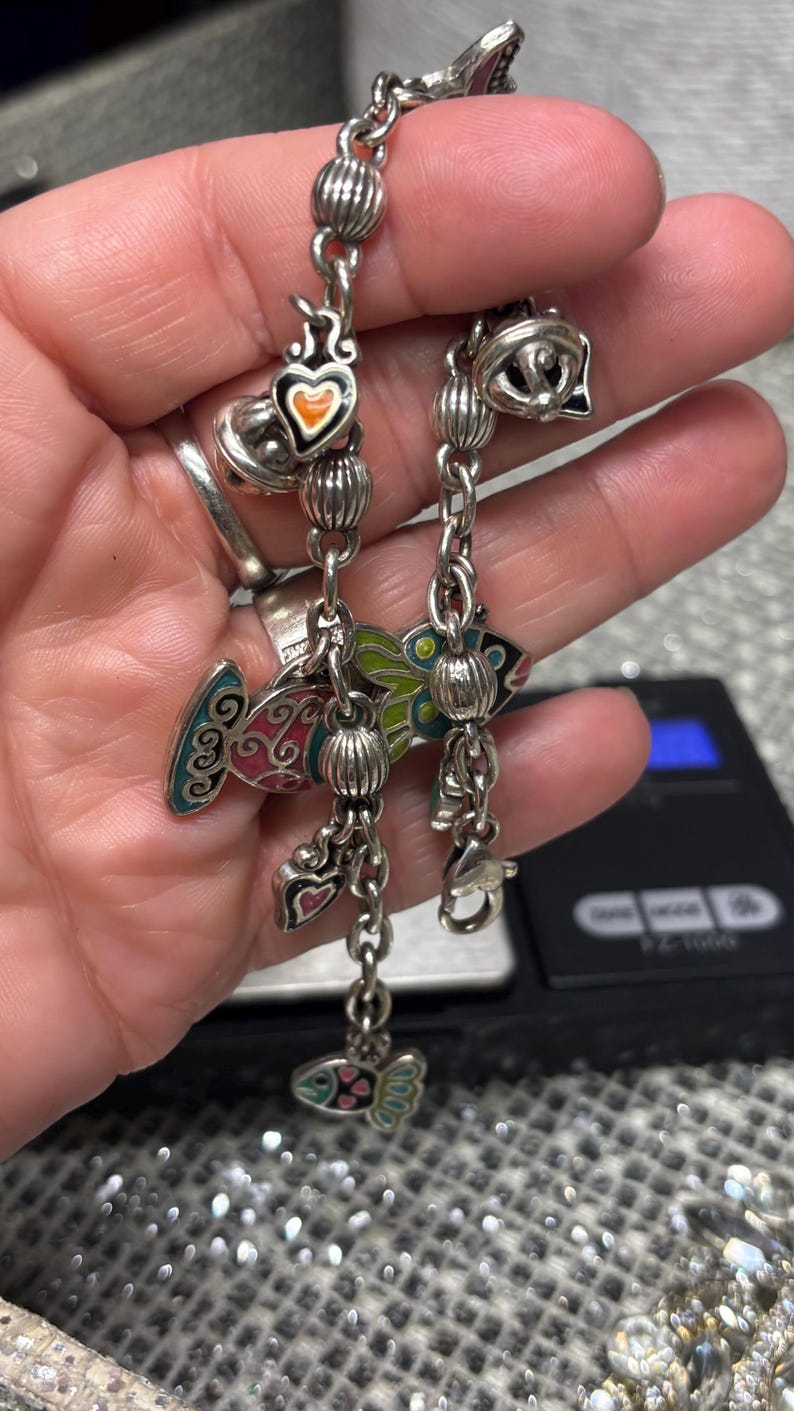 Pu&ograve; includere: Bracciale con ciondoli in argento con pesci in smalto colorato, cuori e perline decorative. Il bracciale ha una chiusura a moschettone ed &egrave; mostrato su uno sfondo sfocato.