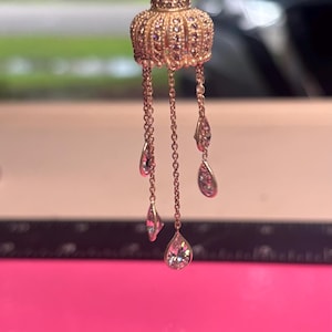 May include: Gold-toned pendant necklace featuring a decorative crown-shaped top adorned with small purple stones. Delicate chains hang from the crown, each suspending a teardrop-shaped clear crystal. The necklace is displayed against a pink background.