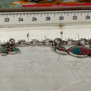 Pu&ograve; includere: Bracciale con ciondoli argentato con vari ciondoli decorativi, tra cui pesci e cuori. I ciondoli sono in pi&ugrave; colori. Un righello &egrave; posto sopra il bracciale, che mostra le misure in centimetri.
