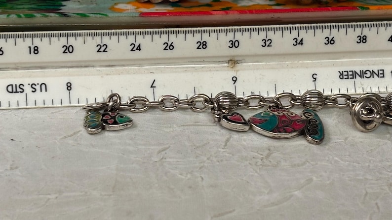 Pu&ograve; includere: Bracciale con ciondoli argentato con vari ciondoli decorativi, tra cui pesci e cuori. I ciondoli sono in pi&ugrave; colori. Un righello &egrave; posto sopra il bracciale, che mostra le misure in centimetri.