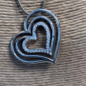 Pode incluir: Colar com pingente de cora&ccedil;&atilde;o prateado. O pingente apresenta tr&ecirc;s cora&ccedil;&otilde;es aninhados, com o cora&ccedil;&atilde;o interior delineado com cristais transparentes. A corrente do colar &eacute; um design delicado e tecido. O fundo &eacute; um material tecido texturizado.