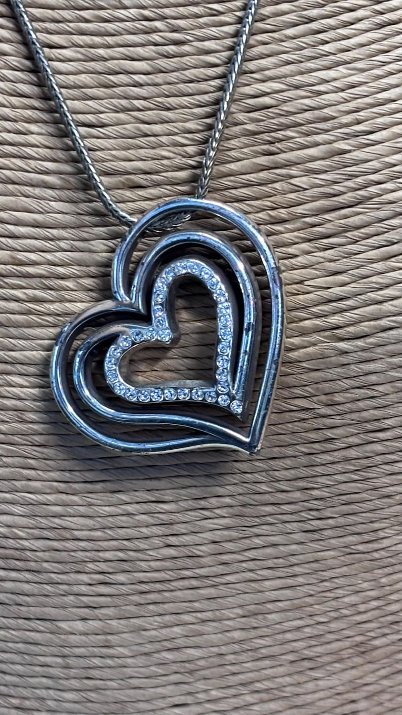 Pode incluir: Colar com pingente de cora&ccedil;&atilde;o prateado. O pingente apresenta tr&ecirc;s cora&ccedil;&otilde;es aninhados, com o cora&ccedil;&atilde;o interior delineado com cristais transparentes. A corrente do colar &eacute; um design delicado e tecido. O fundo &eacute; um material tecido texturizado.