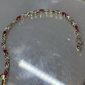 May include: A gold-tone link bracelet featuring a repeating X-shaped pattern set with small clear stones. The bracelet is accented with oval-shaped red gemstones spaced evenly throughout the design and includes a secure metal clasp closure.