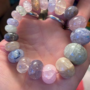 May include: A beaded bracelet featuring a variety of colorful, polished gemstones. The stones are oval and round, in shades of pink, purple, green, and clear. The bracelet is held in a hand.