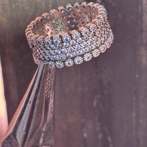 May include: A silver ring with multiple rows of small, clear gemstones. The ring is held by a pair of pliers against a dark, textured background. The ring's design features a continuous band of sparkling stones.