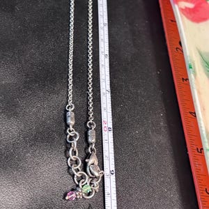 Puede incluir: Collar plateado con una cadena delicada y elementos decorativos. El collar presenta peque&ntilde;as piedras preciosas de colores y un colgante. Una regla muestra que el collar mide aproximadamente 25 cm de largo.