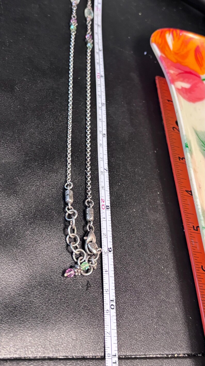 Puede incluir: Collar plateado con una cadena delicada y elementos decorativos. El collar presenta peque&ntilde;as piedras preciosas de colores y un colgante. Una regla muestra que el collar mide aproximadamente 25 cm de largo.