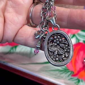 Puede incluir: Collar plateado con un colgante ovalado con un dise&ntilde;o floral y de abeja. El collar tiene m&uacute;ltiples cadenas, cuentas rosas y peque&ntilde;os abalorios. El colgante mide aproximadamente 3,8 cm de largo.