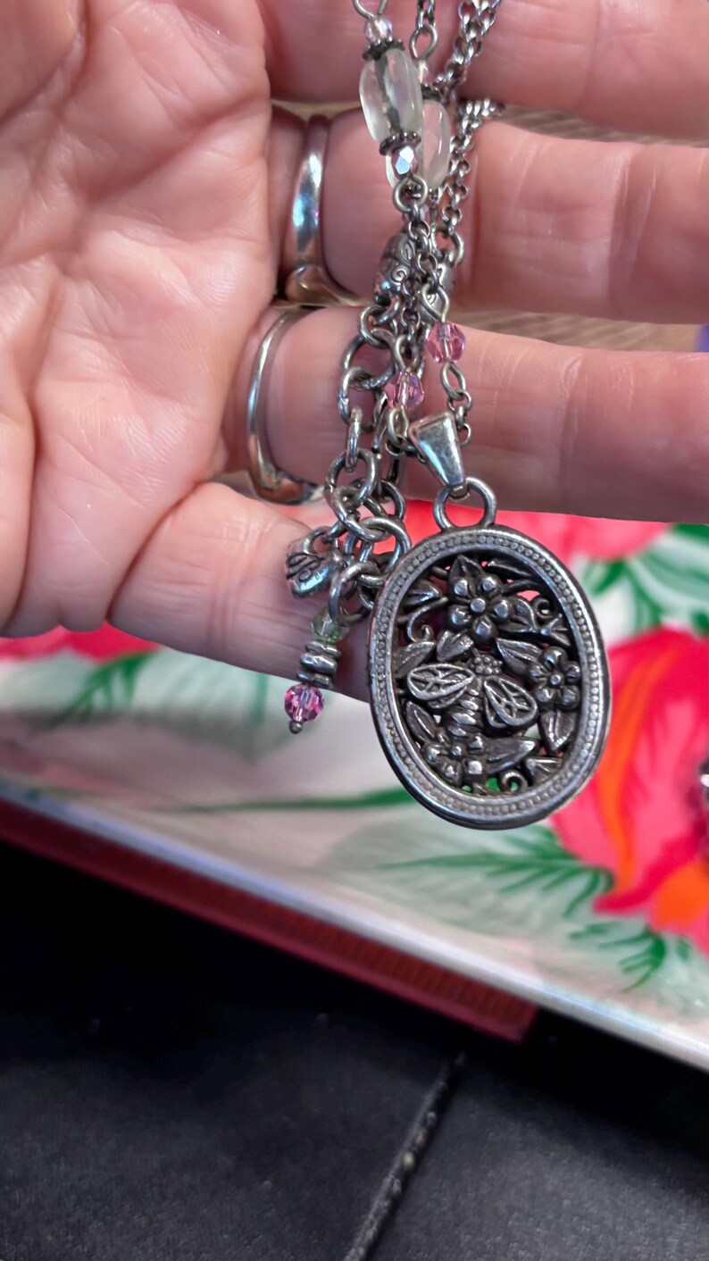 Puede incluir: Collar plateado con un colgante ovalado con un dise&ntilde;o floral y de abeja. El collar tiene m&uacute;ltiples cadenas, cuentas rosas y peque&ntilde;os abalorios. El colgante mide aproximadamente 3,8 cm de largo.