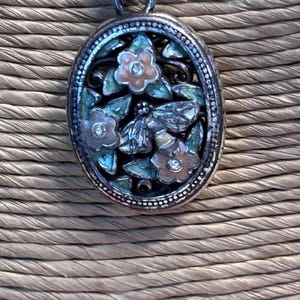 Puede incluir: Collar con colgante plateado con dise&ntilde;o floral ovalado. El colgante presenta una abeja, flores y hojas en tonos rosa, verde y plateado. El collar tiene una cadena delicada y detalles de cuentas rosas.