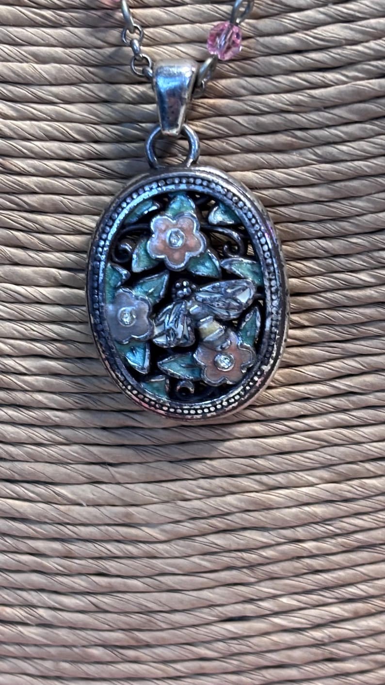 Puede incluir: Collar con colgante plateado con dise&ntilde;o floral ovalado. El colgante presenta una abeja, flores y hojas en tonos rosa, verde y plateado. El collar tiene una cadena delicada y detalles de cuentas rosas.