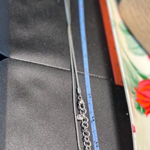 Pode incluir: Um colar de prata com corrente e um pingente em forma de cora&ccedil;&atilde;o. O colar &eacute; medido com uma r&eacute;gua azul, mostrando um comprimento de aproximadamente 40 cm. O colar est&aacute; em uma superf&iacute;cie preta.