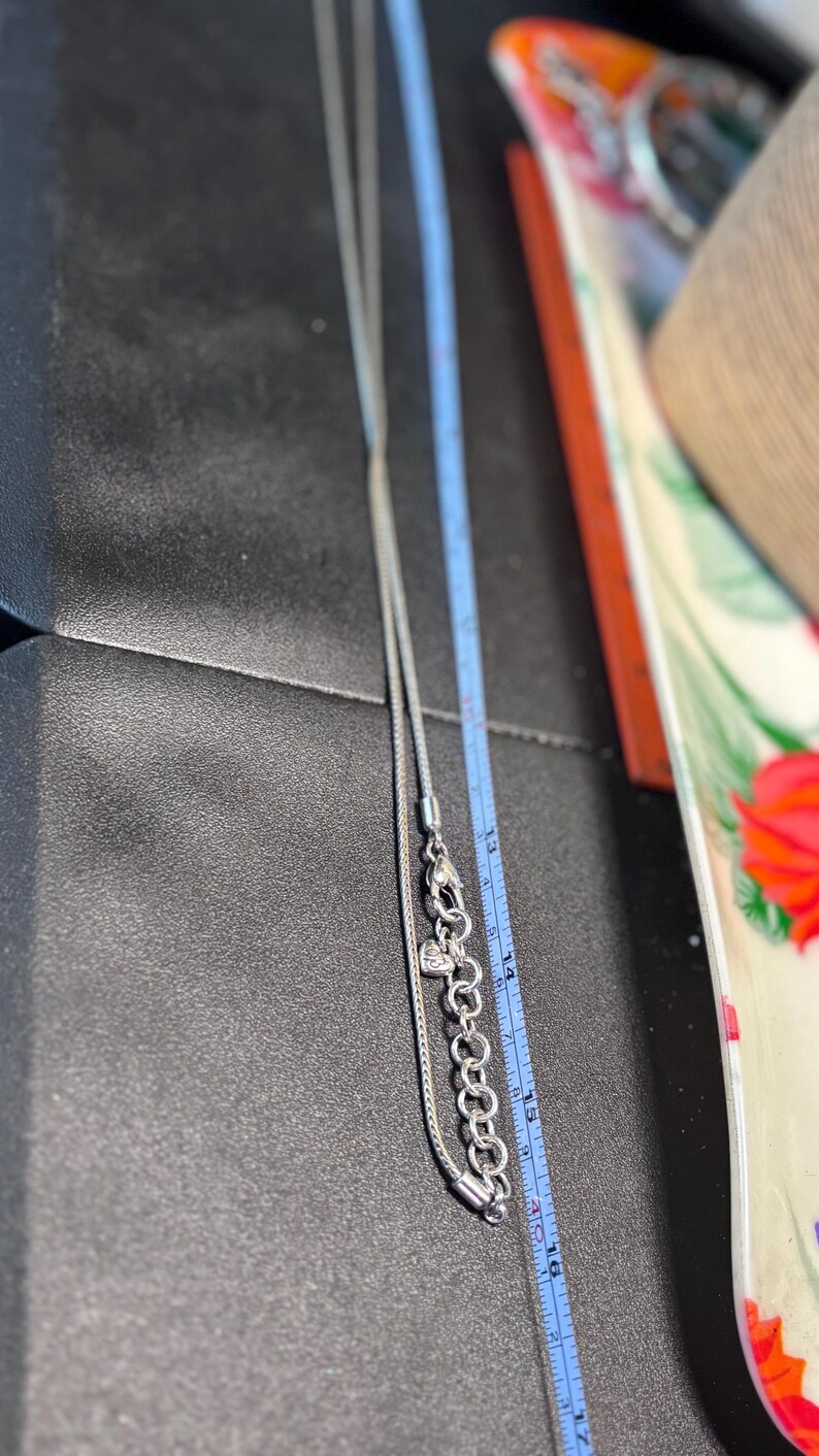 Pode incluir: Um colar de prata com corrente e um pingente em forma de cora&ccedil;&atilde;o. O colar &eacute; medido com uma r&eacute;gua azul, mostrando um comprimento de aproximadamente 40 cm. O colar est&aacute; em uma superf&iacute;cie preta.