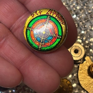 May include: A round, gold-toned pin with the text "Def Leppard" in a gold font. The pin features a target design with concentric circles in green, red, blue, and gold. Additional gold-toned pins are visible.