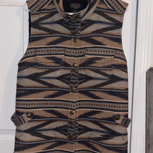May include: A sleeveless vest with a geometric pattern in shades of brown, gray, and blue. The vest has a high collar, a button-up front, and two front pockets. The vest is hanging on a white hanger.