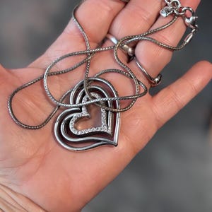 Pode incluir: Colar prateado com um pendente de cora&ccedil;&atilde;o em camadas. O pendente tem tr&ecirc;s formas de cora&ccedil;&atilde;o, com o cora&ccedil;&atilde;o mais interno adornado com pequenos cristais transparentes. O colar &eacute; exibido em uma m&atilde;o, mostrando a delicada corrente e o design do pendente.