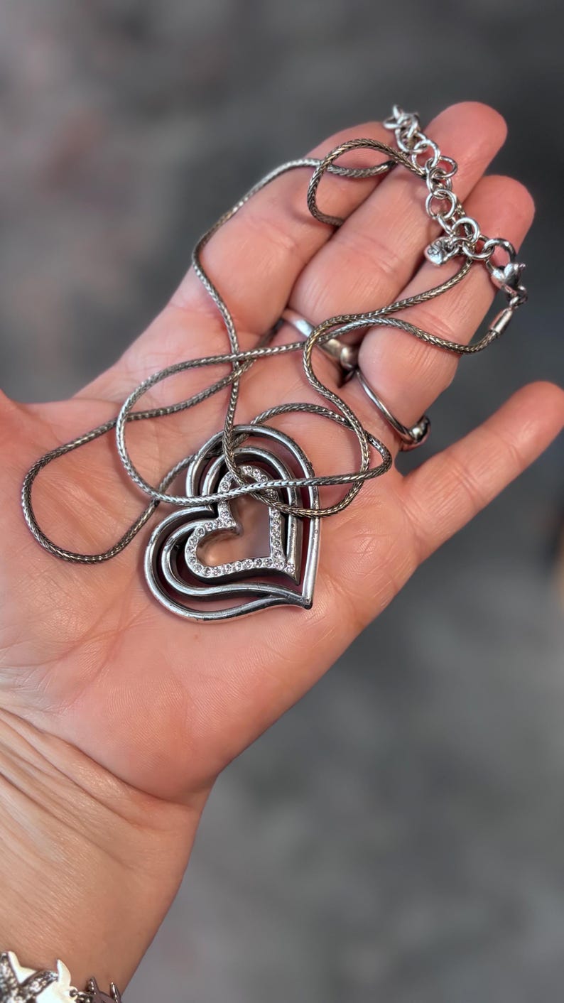 Pode incluir: Colar prateado com um pendente de cora&ccedil;&atilde;o em camadas. O pendente tem tr&ecirc;s formas de cora&ccedil;&atilde;o, com o cora&ccedil;&atilde;o mais interno adornado com pequenos cristais transparentes. O colar &eacute; exibido em uma m&atilde;o, mostrando a delicada corrente e o design do pendente.